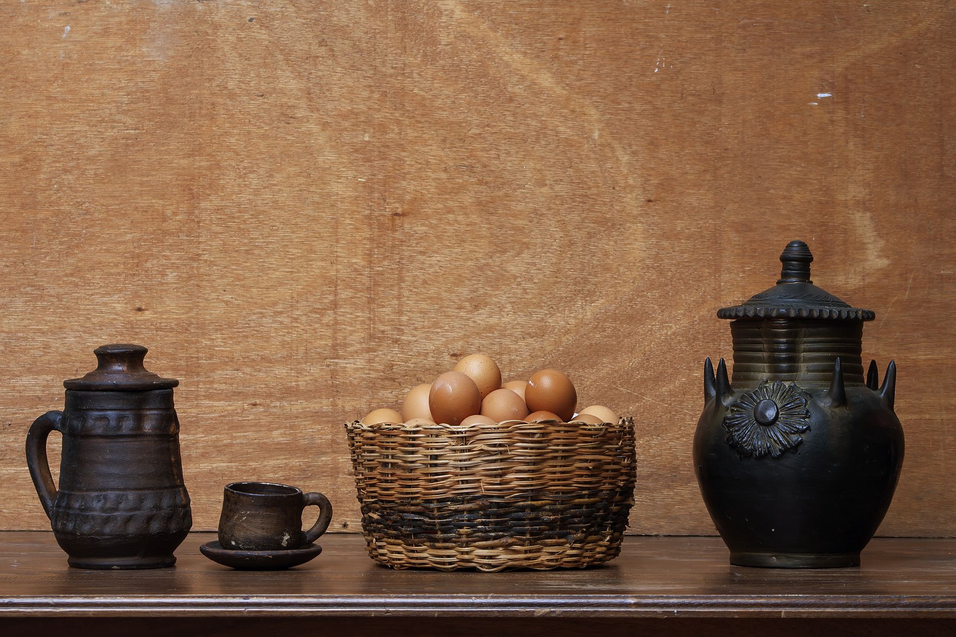 A rustic tabletop setup features pottery, a wicker basket with eggs, and antique jugs.